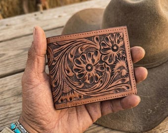 Pode incluir: Uma carteira de couro castanho-claro com padrões florais intrincados e volutas gravados na superfície frontal. A carteira tem uma forma retangular com bordas costuradas e um acabamento natural em castanho-claro com detalhes em castanho-escuro.