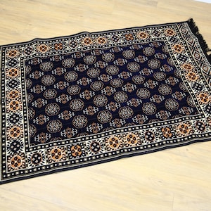 May include: A black rug with a detailed pattern of orange, white, and blue geometric shapes. The rug has a fringe border.