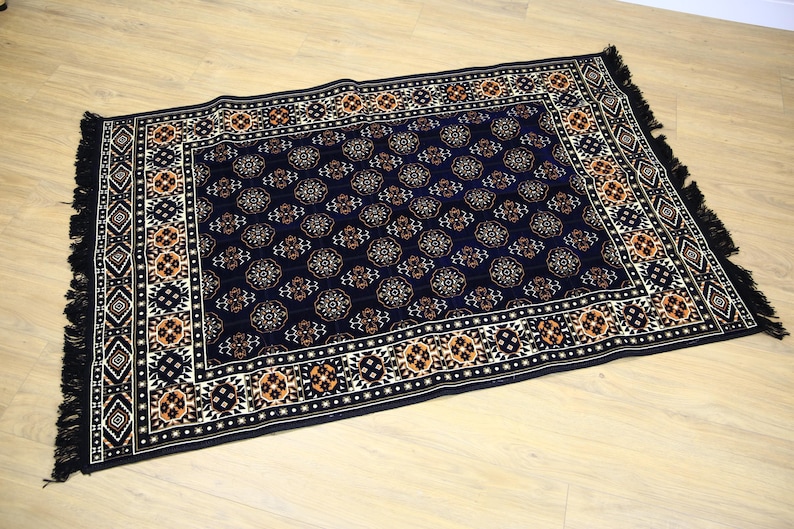 May include: A black rug with a detailed pattern of orange, white, and blue geometric shapes. The rug has a fringe border.