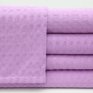 May include: A stack of four lavender-colored waffle weave towels. The towels are folded and stacked on a white surface.