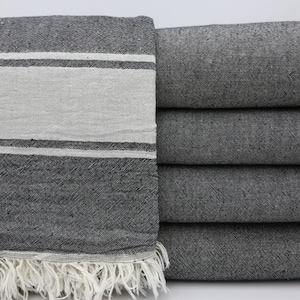 Può includere: Una coperta a righe bianche e nere con un bordo a frangia. La coperta è piegata e impilata su una superficie bianca.