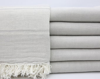 May include: A stack of light gray cotton throw blankets with white fringe. The blankets are folded and stacked on top of each other.