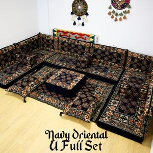 Op de afbeelding: Een U-vormige vloerzitset in marineblauw en goud met ingewikkelde patronen. De set bevat meerdere kussens en een vierkante ottomane. De set is in een woonkamer met een houten vloer gerangschikt.