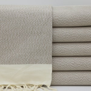 May include: A stack of six beige Turkish towels with a white border and fringe. The towels are made of cotton and have a textured weave.