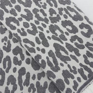 May include: A white fabric with a gray leopard print pattern. The fabric has a fringed edge.
