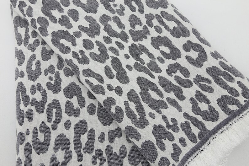 May include: A white fabric with a gray leopard print pattern. The fabric has a fringed edge.