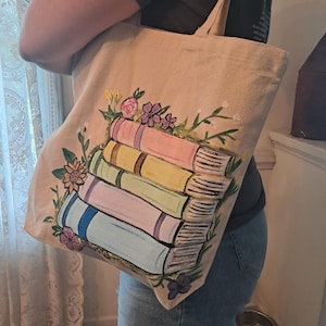 Puede incluir: Bolso tote de lona beige con una pila de libros coloridos y detalles florales. Los libros están pintados en tonos pastel, como rosa, amarillo y azul. El bolso tiene dos asas y un diseño sencillo.