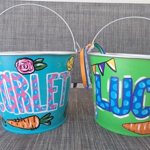 May include: Two hand-painted metal buckets with white handles. One is teal with the name "Scarlet" in pink and blue polka dots, a carrot, and floral accents. The other is green with "Luca" and a carrot.