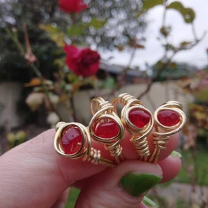 Puede incluir: Cuatro anillos de alambre dorado envueltos con acentos de piedras preciosas rojas. Los anillos están apilados en un dedo.