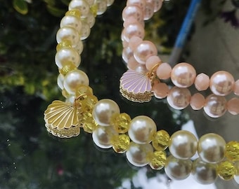May include: Two pearl necklaces with gold shell charms. One necklace is made of white pearls with yellow accents, and the other is made of pink pearls.