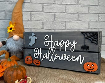 May include: A gray wooden crate with the words "Happy Halloween" in white letters. The crate is decorated with a ghost, a spider web, and a spider. There are also several small orange pumpkins with black faces.