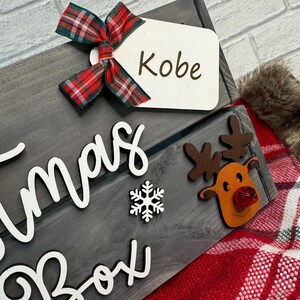 May include: A gray wooden Christmas box with a white snowflake and the words "Christmas Box" written on it. A red and black plaid bow with a white tag that says "Kobe" is attached to the top of the box. A brown reindeer with a red nose is attached to the side of the box.