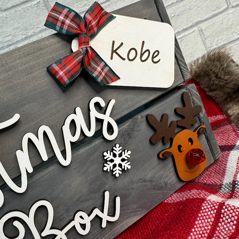 May include: A gray wooden Christmas box with a white snowflake and the words "Christmas Box" written on it. A red and black plaid bow with a white tag that says "Kobe" is attached to the top of the box. A brown reindeer with a red nose is attached to the side of the box.
