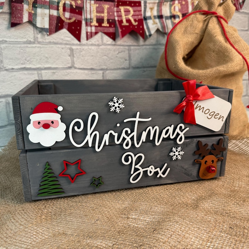 May include: A gray wooden crate with the words "Christmas Box" written on it. The crate is decorated with a Santa Claus face, a reindeer, snowflakes, and stars. There is a red ribbon tied around the crate with a tag that says "mogen".