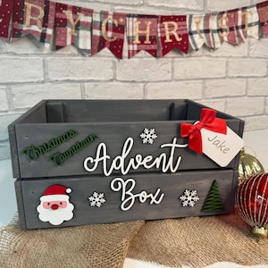 May include: A grey wooden crate with the words "Christmas Countdown" and "Advent Box" on the front. The crate is decorated with snowflakes, a Christmas tree, and a Santa Claus head. A red ribbon and a white tag with the name "Jake" are attached to the crate.