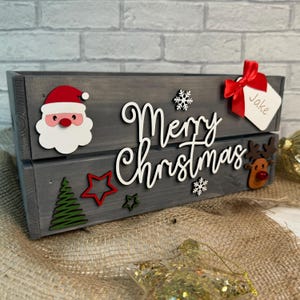 May include: A grey wooden crate with a white "Merry Christmas" sign, red and green Christmas decorations, and a white tag with the name "Jake" written on it.