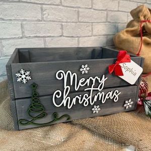 May include: A grey wooden Christmas crate with a white "Merry Christmas" sign, a green Christmas tree, and white snowflakes. The crate has a red ribbon and a white tag with the name "Sophia" written on it.