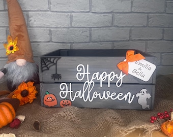 May include: A gray wooden Halloween-themed box with the words "Happy Halloween" in white script. The box is decorated with a spiderweb, spider, pumpkins, and a ghost. An orange bow and tag with the names "Emilia & Bella" are attached.