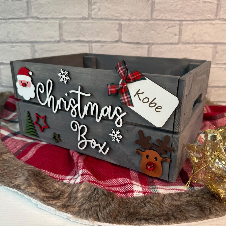 May include: A gray wooden Christmas box with a red and black plaid bow, a white tag with the name "Kobe" written on it, and various Christmas decorations including a Santa Claus, a snowflake, a star, and a reindeer. The box is labeled "Christmas Box".