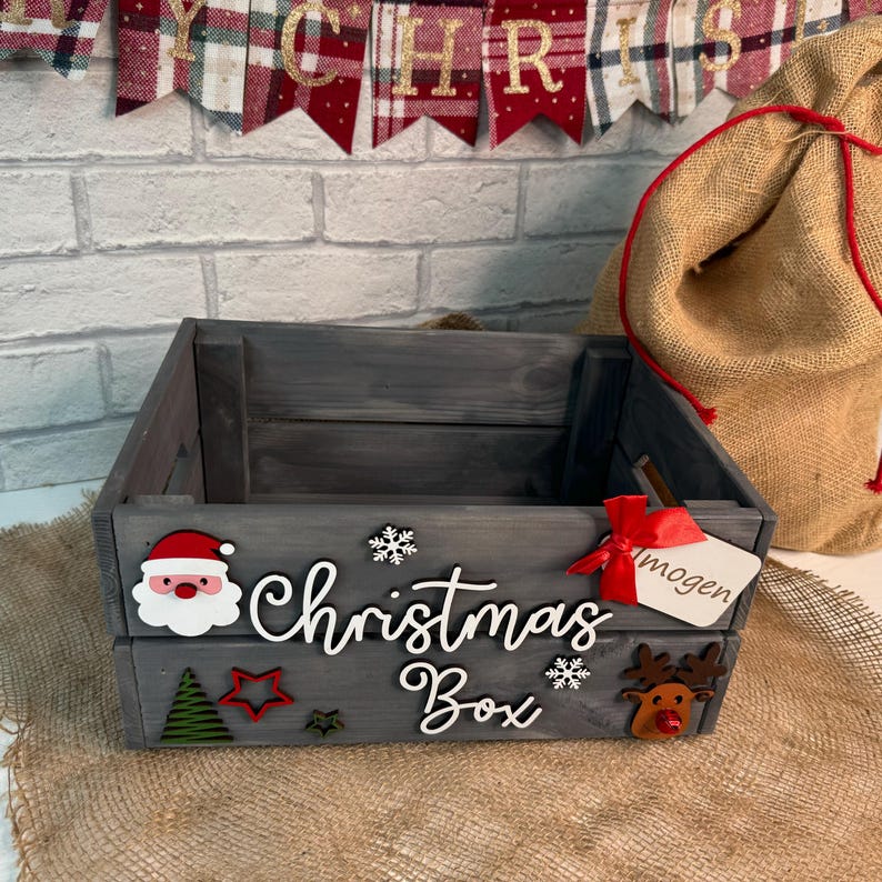 May include: A gray wooden Christmas box with a red ribbon and a tag that says "mogen". The box is decorated with a Santa Claus, a snowflake, a Christmas tree, a star, and a reindeer.