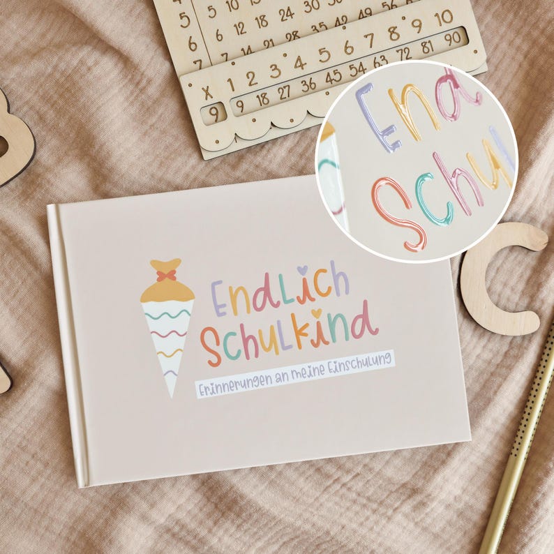 K&ouml;nnte beinhalten: Ein hellbeiges, rechteckiges Buch mit einer Grafik einer bunten Schult&uuml;te und dem Text Endlich Schulkind sowie Erinnerungen an meine Einschulung. Das Cover zeigt pastellfarbene Schriftz&uuml;ge und ein kleines Herz-Symbol in einem klaren Design.