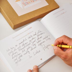 K&ouml;nnte beinhalten: Ein offenes Tagebuch mit linierten Seiten und handschriftlichem Text in schwarzer Tinte, betitelt mit Pers&ouml;nliche Worte meiner Eltern. Eine Hand h&auml;lt einen gelben Stift und schreibt auf die Seite. Ein brauner Karton liegt oberhalb des Buches.