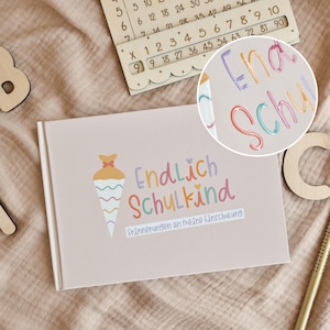 Könnte beinhalten: Ein hellrosa rechteckiges Buch mit einer Grafik einer bunten Schultüte und dem Text Endlich Schulkind Erinnerungen an meine Einschulung. Das Cover zeigt pastellfarbene Schriftzüge in Lila, Orange und Rosa sowie ein kleines Herzsymbol.