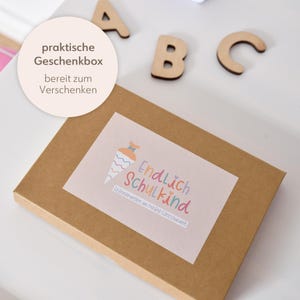 Peut inclure: Une bo&icirc;te cadeau rectangulaire en carton brun avec une &eacute;tiquette illustr&eacute;e d'un cornet d'&eacute;cole color&eacute; et le texte Endlich Schulkind et Erinnerungen an meine Einschulung. Des lettres en bois A, B et C sont plac&eacute;es pr&egrave;s de la bo&icirc;te.