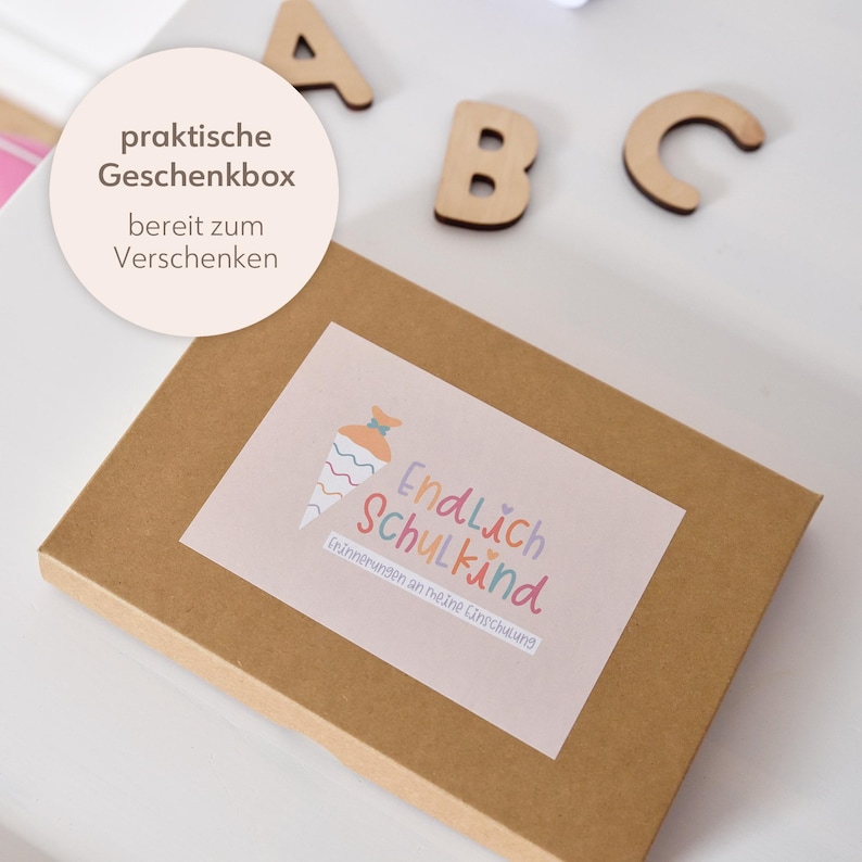 Peut inclure: Une bo&icirc;te cadeau rectangulaire en carton brun avec une &eacute;tiquette illustr&eacute;e d'un cornet d'&eacute;cole color&eacute; et le texte Endlich Schulkind et Erinnerungen an meine Einschulung. Des lettres en bois A, B et C sont plac&eacute;es pr&egrave;s de la bo&icirc;te.