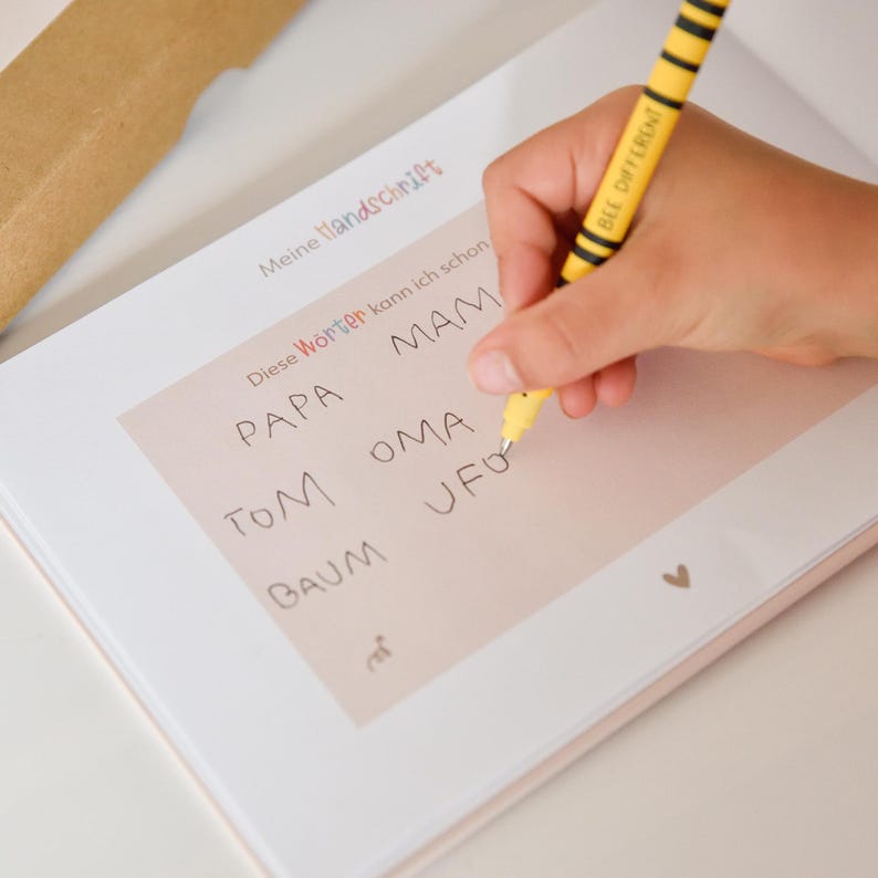 K&ouml;nnte beinhalten: Ein wei&szlig;es &Uuml;bungsheft mit dem aufgedruckten Text Meine Handschrift und Diese W&ouml;rter kann ich schon. Die Seite enth&auml;lt handschriftliche W&ouml;rter wie PAPA, MAM, TOM, OMA, BAUM und UFO. Ein gelb-schwarz gestreifter Stift wird zum Schreiben verwendet.