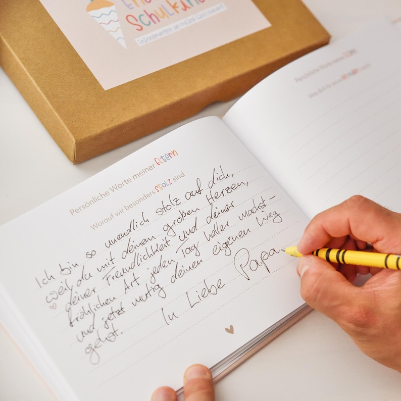 Peut inclure: Un journal ouvert avec des pages lign&eacute;es contenant un texte manuscrit en allemand &agrave; l'encre noire, intitul&eacute; Pers&ouml;nliche Worte meiner Eltern. Une main tient un crayon jaune et &eacute;crit sur la page. Une bo&icirc;te en carton brun est plac&eacute;e au-dessus.