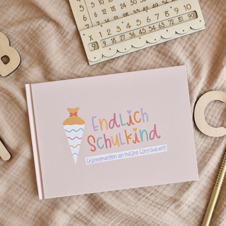 Peut inclure: Un livre rectangulaire rose clair avec un graphique de c&ocirc;ne color&eacute; et le texte Endlich Schulkind Erinnerungen an meine Einschulung. Le livre est entour&eacute; de lettres en bois et d'une table de multiplication en bois sur un tissu beige.