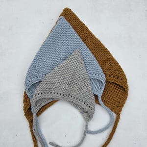 May include: Three knitted baby bonnets in shades of gray, blue, and brown are arranged on a white surface. Each bonnet has a triangular shape with ties. The bonnets are made of a soft, textured yarn.