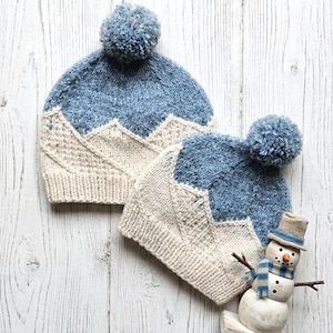Puede incluir: Dos gorros de punto con una sección superior azul y una inferior color crema con un patrón de montañas. Cada gorro tiene un pompón azul en la parte superior. Una pequeña figura decorativa de muñeco de nieve lleva una bufanda azul y crema.