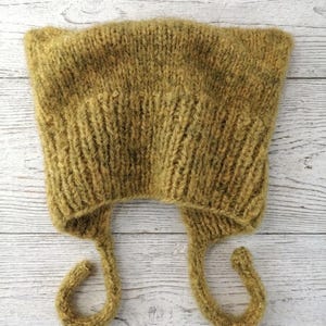 May include: A fuzzy, olive-green knitted bonnet featuring a ribbed texture throughout the main body and two long, thin braided ties extending from the bottom corners. The hat has a soft, brushed appearance with a distinct, slightly pointed shape.