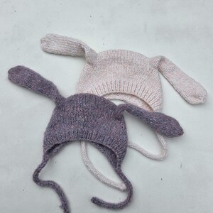 May include: Two knitted bunny hats. One is a light purple colour, and the other is a light pink colour. Both hats have long, floppy ears and ties under the chin. The hats are made of a soft, textured yarn.