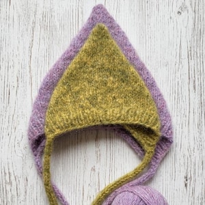 Könnte beinhalten: Eine gestrickte Haube mit spitzer Oberseite aus texturiertem, flauschigem Garn. Die Innenschicht ist in Olivgrün gehalten, während die Außenschicht in einem sanften Lavendelton erscheint. Die Haube verfügt über lange, passende Strickbänder.