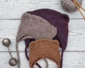 May include: Three knitted bonnets with small ear-like protrusions at the top, featuring long chin ties. The bonnets are colored in deep purple, light brown, and tan. A ball of grey yarn with two wooden knitting needles and a silver rattle are included.