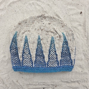 Op de afbeelding: Een gebreide muts met een gespikkelde haverkleurige basis en een reeks van vijf driehoekige blauwe patronen die vanaf de onderrand omhoog lopen. De muts is afgewerkt met een effen blauwe boord in ribbelsteek langs de onderkant.