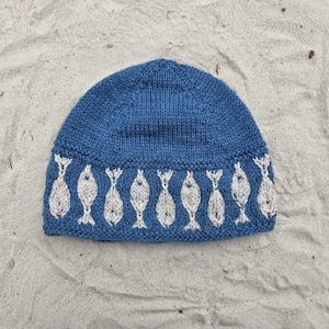 May include: A blue knit beanie featuring a repeating pattern of light gray fish motifs around the lower band. The hat is constructed with a standard stockinette stitch on the crown and a ribbed edge at the base, showcasing a textured yarn finish.