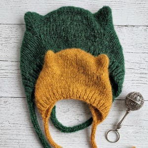 May include: Two knitted bonnets with ear-like protrusions, one in dark forest green and one in mustard yellow, featuring long chin ties. Beside the bonnets lies a small, round, silver-toned metal rattle with an intricate filigree pattern.