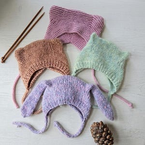 Puede incluir: Cuatro gorros tejidos a mano con detalles inspirados en animales, incluyendo dos con orejas puntiagudas en malva y verde menta, uno con orejas redondeadas en color canela y otro con largas orejas de conejo en lavanda moteado, junto a dos agujas.