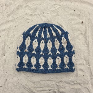 May include: A knitted beanie hat featuring a deep blue base colour with a repeating pattern of light beige fish motifs arranged in horizontal rows. The hat has a rounded crown and a ribbed edge, showcasing a textured yarn construction throughout the design.