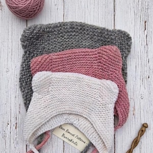 Op de afbeelding: Drie gebreide mutsjes met berenoortjes in grijs, oudroze en crème, verticaal gestapeld. Een bol bijpassend roze garen ligt linksboven en een houten breinaald rechtsonder. Een label bevat de tekst Bear Bonnet Patterns BurraKnits.