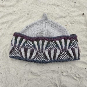 May include: A light grey knit beanie featuring a pointed crown and a wide, folded brim. The brim displays a decorative pattern of vertical, fan-shaped motifs in shades of dark purple, green, and grey, set against a textured, speckled background.