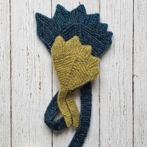 Puede incluir: Un gorro de punto que presenta una gran cresta en forma de abanico de color azul cerceta oscuro con bordes puntiagudos, superpuesta a una pieza más pequeña de color verde chartreuse texturizado, con largos lazos de punto azul cerceta.