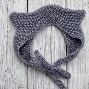 Puede incluir: Un gorro tejido de color gris oscuro con textura de punto bobo y dos formas puntiagudas en la parte superior que recuerdan a unas orejas. La prenda incluye dos lazos tejidos integrados en la base para atar bajo la barbilla.