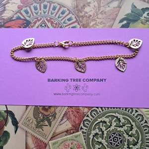 Peut inclure: Bracelet en chaîne dorée avec des breloques en forme de feuilles. Le bracelet présente un design de chaîne délicat avec plusieurs pendentifs en forme de feuilles. Il est présenté sur une surface violette avec le texte "BARKING TREE COMPANY".