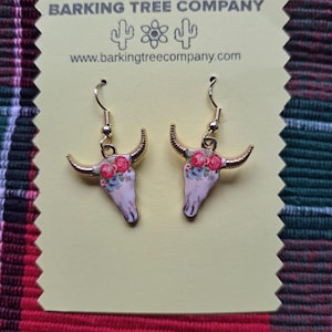 Peut inclure: Boucles d'oreilles dorées avec un motif de crâne blanc et des accents floraux. Le crâne est orné d'une couronne florale dans des tons de rouge, rose et bleu. Les boucles d'oreilles sont étiquetées "Barking Tree Company" et "www.barkingtreecompany.com".