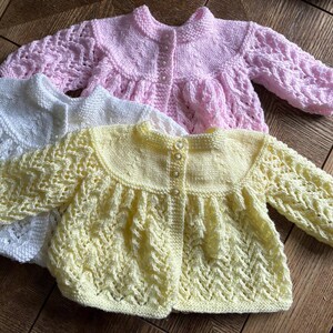 May include: Three hand-knitted baby cardigans in pastel colors: white, pink, and yellow. Each cardigan features a round collar, button closure, and intricate openwork sleeves. The garments are made of soft yarn, perfect for a baby.